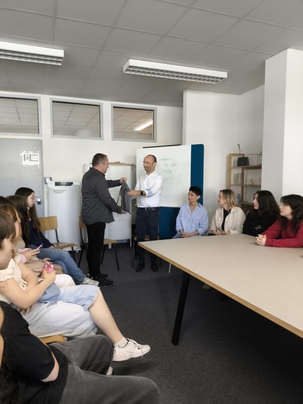 Zajęcia warsztatowe – dwóch prowadzących przy tablicy flipchart, uczniowie po obu stronach stołu konferencyjnego