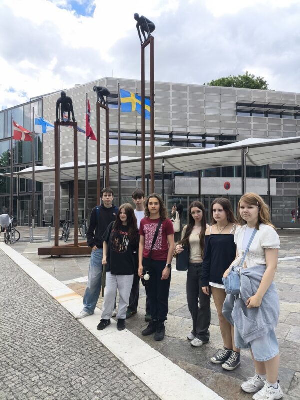Uczniowie TME przed siedzibą organizacji młodzieżowej z rzeźbami wspinaczek na słupach i flagami europejskich krajów
